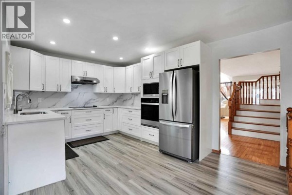 Contemporary white kitchen with stainless steel appliances, light wood floors, and view of staircase and living area. - 16 Sun Harbour Crescent Se, Calgary