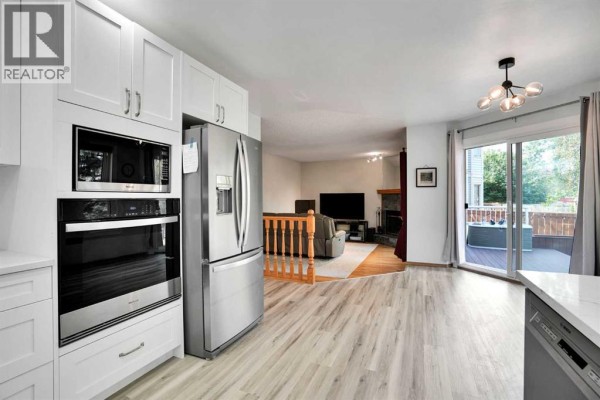 Wide view of a white kitchen with stainless steel appliances, opening to a living area with fireplace and patio doors. - 16 Sun Harbour Crescent Se, Calgary
