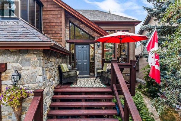 Close-up view of the front entrance with wooden steps, wicker chairs, a red umbrella, and a Canadian flag. - 160 Carey, Canmore