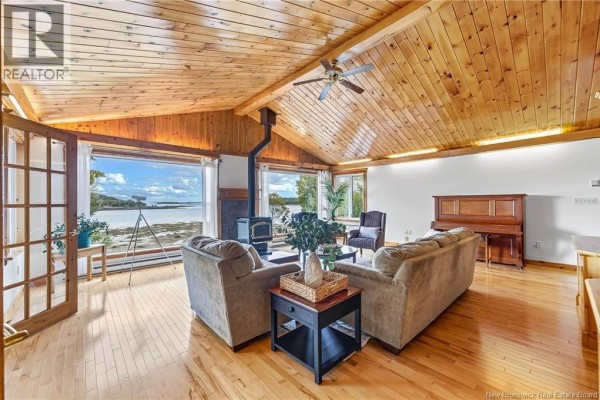 Living room with wood-paneled vaulted ceiling, wood floors, fireplace, and large windows overlooking water. - 160 Harkness Road, Chamcook