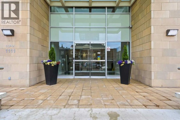 Entrance to the apartment building with glass doors, stone facade, and decorative planters. - 1602, 1111 10 Street Sw, Calgary