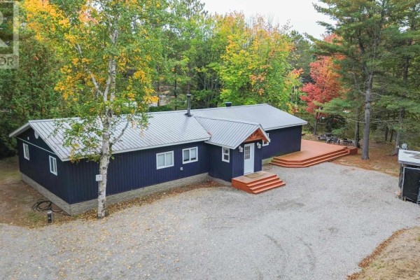 Exterior view of a dark blue bungalow with a silver metal roof, large gravel driveway and surrounding forest. - 1602 Pineshores Rd, Goulais River