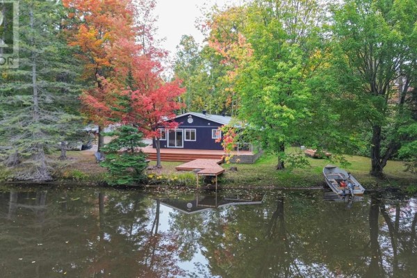 View of a dark blue bungalow with a wooden deck and dock from across a calm body of water. - 1602 Pineshores Rd, Goulais River
