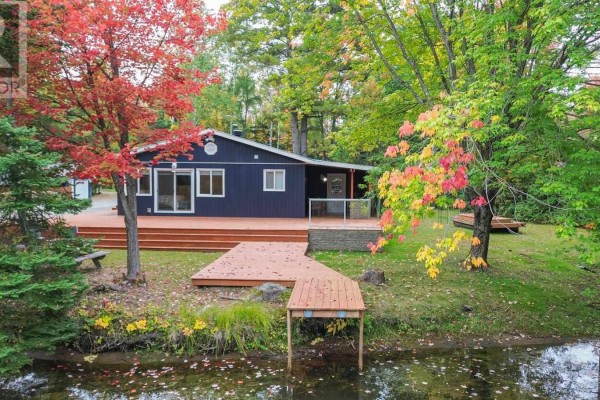 A dark blue bungalow with multiple decks and a small dock extending into the water during autumn. - 1602 Pineshores Rd, Goulais River
