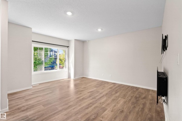 Empty living room with a large window, wood flooring, and a mounted TV. - 16040 11 Av Sw, Edmonton