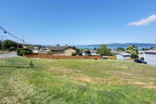 Grassy lot with a wooden fence, houses, trees, and a distant water view. - 1605 Adelaide St, Crofton