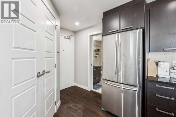 Kitchen area with dark cabinetry, stainless steel refrigerator, and hallway leading to a bathroom. - 1610, 225 11 Avenue Se, Calgary