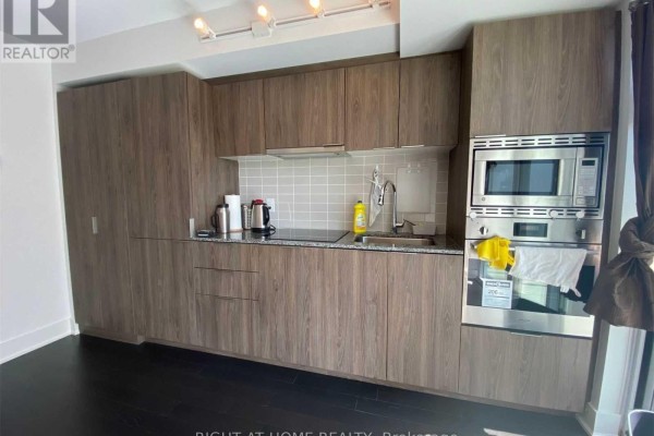 Modern kitchen with wood-grain cabinets, stainless steel appliances, and light grey backsplash. - 1612 - 955 Bay Street, Toronto