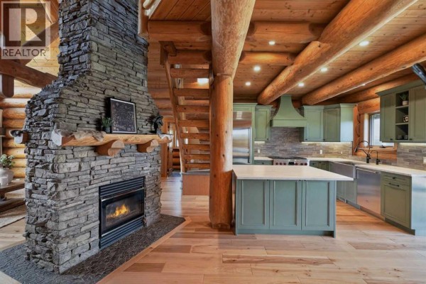 Interior great room with a large stone fireplace, wood beams, and a green kitchen. - 162104 376 Street W, Rural Foothills County