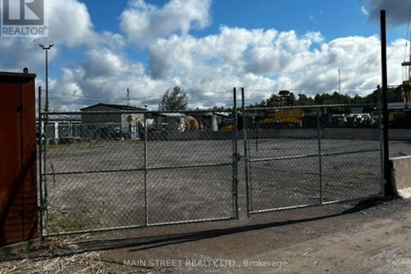 Double chain-link gate with barbed wire topping, leading into a gravel yard with industrial structures beyond. - 163 Ram Forest Road, Whitchurch Stouffville