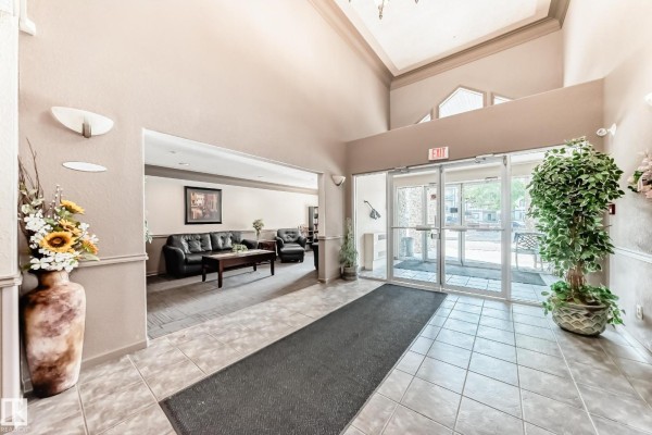 Apartment building lobby with seating area, potted plants, and double glass doors leading outside. - #138 16311 95 St Nw, Edmonton