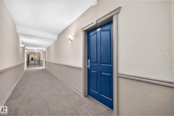 Interior hallway of an apartment building with beige walls, carpeted floors, and a blue suite door. - #138 16311 95 St Nw, Edmonton