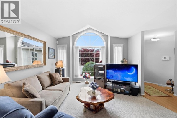 Living room with a beige sofa, large mirror, entertainment center, and a large arched window. - 1634 Carmi Avenue Unit# 121, Penticton