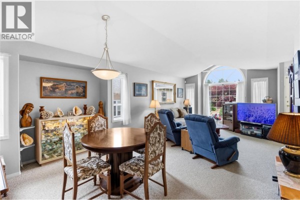 Dining area with a round wooden table, upholstered chairs, and a view into the living room. - 1634 Carmi Avenue Unit# 121, Penticton