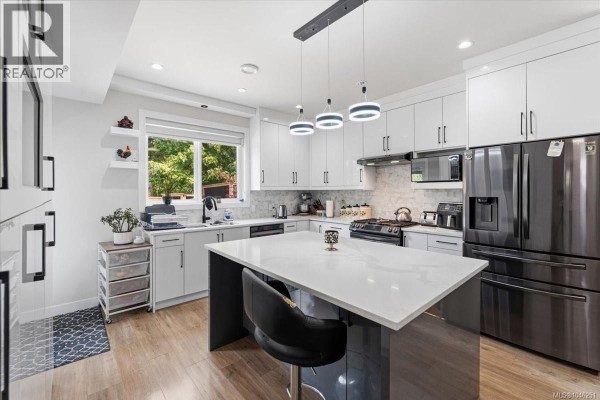 White kitchen with a large island, black appliances, and light wood flooring. - 1636 Roberta Rd S, Nanaimo