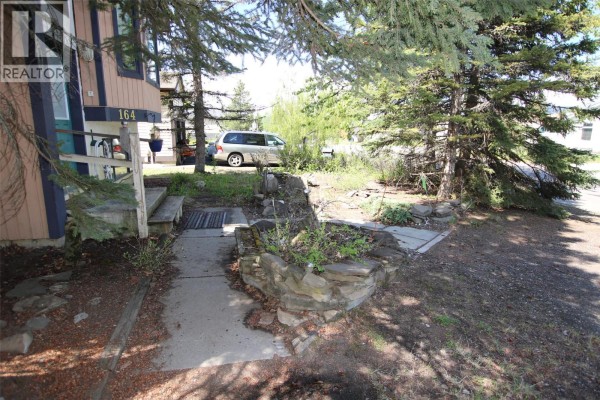 Front entrance of a house with a walkway, trees, and a parked car. - 164 Bergeron Drive, Tumbler Ridge