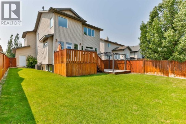 Backyard view of a house with a wooden deck, green grass, and a fence. - 164 Rivermill Landing W, Lethbridge