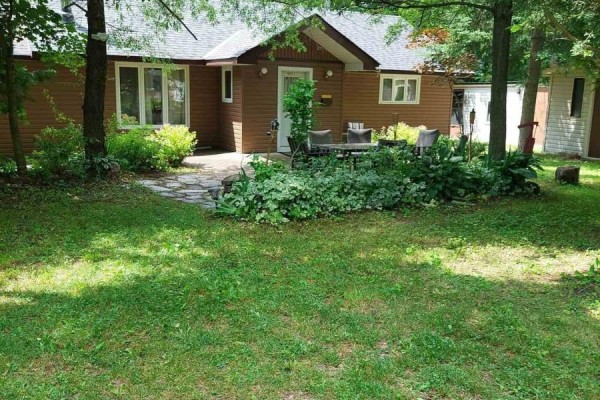 Side view of a brown wood-sided bungalow with a stone walkway and lush green lawn. - 16431 Hwy 35, Algonquin Highlands