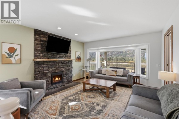 Living room with a stone fireplace, grey sofas, a coffee table, and a large bay window view. - 165 Hoovers Ferry Road, Kamloops