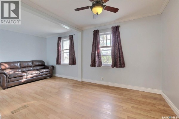 Living room with light blue walls, light wood floors, a brown leather sofa, and two windows with patterned curtains. - 1659 Montreal Street, Regina