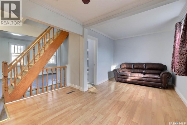 Living area with light wood floors, light blue walls, a brown leather sofa, and a wooden staircase leading upstairs. - 1659 Montreal Street, Regina