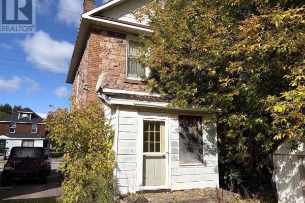 Backyard view with patchy grass, stone patio, large trees, and a brown fence. - 166 Hudson St, Sault Ste  Marie