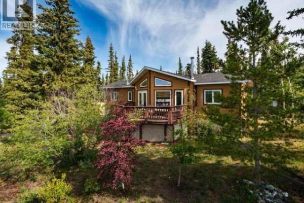 Front view of a wood-sided house with a large deck, nestled amongst mature pine trees. - 166 Judas Creek Drive, Whitehorse South