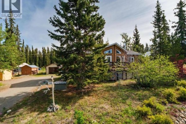 Street view of a property with a brown wood house and deck, surrounded by trees and vegetation. - 166 Judas Creek Drive, Whitehorse South