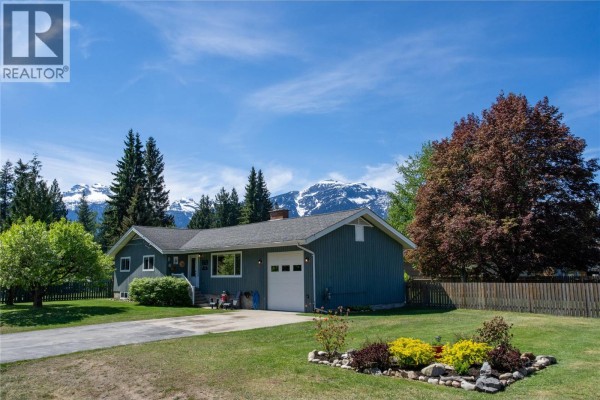 A wide-angle view of the grey house from the driveway, showing the lawn and trees. - 1669 Hiob Road, Revelstoke