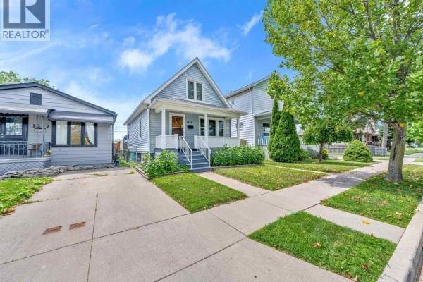 Front view of a light blue house with a driveway, sidewalk, and mature trees. - 1673 Cadillac Street, Windsor
