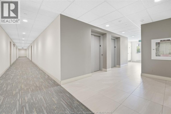 Long apartment building hallway with beige walls, grey carpet, tile floor, and elevator doors. - 1675 Banwell Road Unit# 404, Windsor