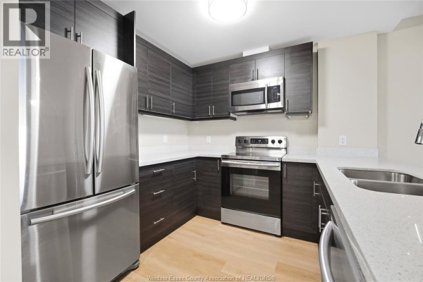 Modern kitchen with dark wood cabinets, stainless steel appliances, white countertops, and light wood floors. - 1675 Banwell Road Unit# 404, Windsor