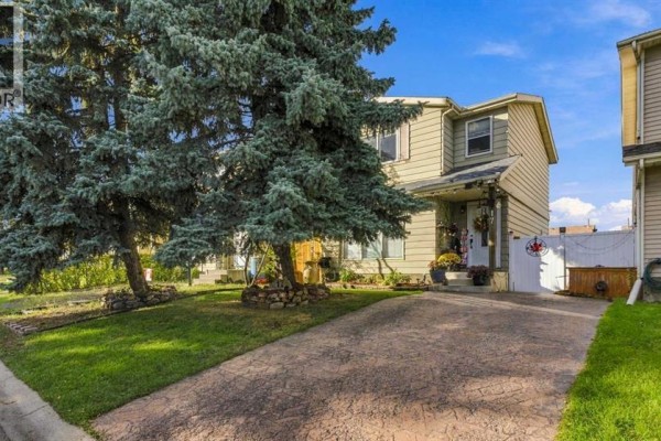 Side exterior view of a two-story house with beige siding and large green evergreen trees. - 17 Erin Ridge Road Se, Calgary
