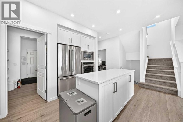 Bright white kitchen with a large island, stainless steel refrigerator, and a staircase with carpeted treads. - 17 Masters Terrace Se, Calgary