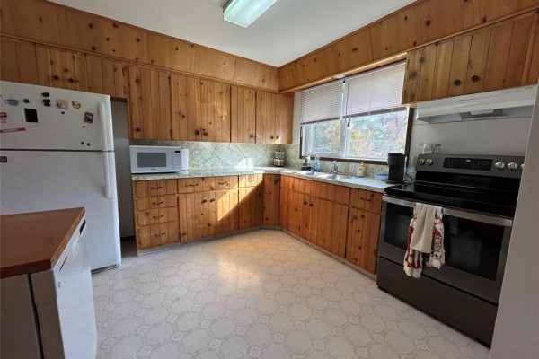 A U-shaped kitchen with wood-paneled walls, wooden cabinets, white appliances, and a window overlooking greenery. - 170 7th Avenue Sw, Minnedosa