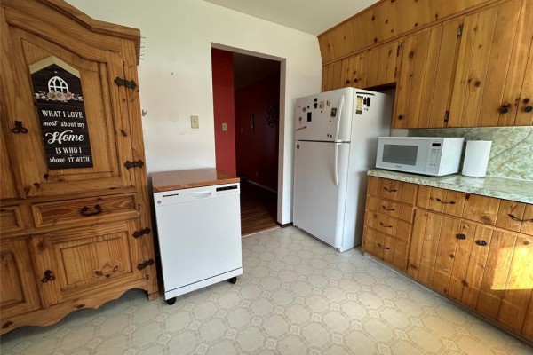 Kitchen view showing a dishwasher, refrigerator, microwave, and wood cabinets, with a doorway to a red hallway. - 170 7th Avenue Sw, Minnedosa
