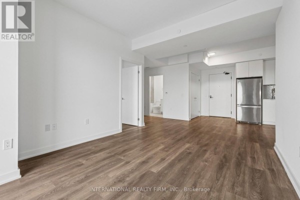 Empty apartment living space with hardwood floors, white walls, and stainless steel refrigerator. - 1701 - 1606 Charles Street, Whitby