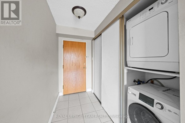 Apartment entryway with a light wood door, tile floor, and stacked washer/dryer. - 1705 - 199 Kent Street, Ottawa