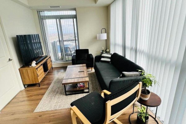 Living room with black sofa, wooden coffee table, television, and floor-to-ceiling windows with vertical blinds. - 1706 - 349 Rathburn Road W, Mississauga