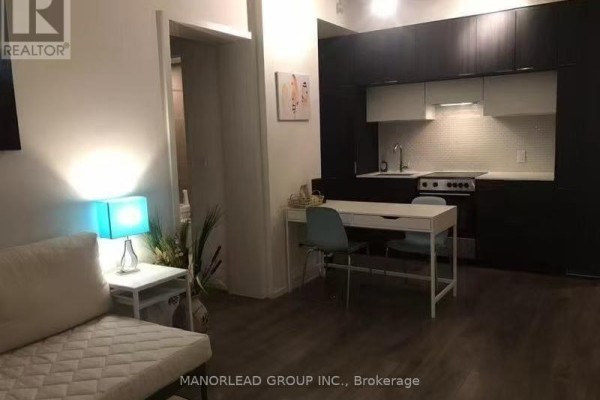 A studio apartment living area with a sofa, desk, and an open concept kitchen with dark cabinetry. - 1708 - 159 Dundas Street E, Toronto