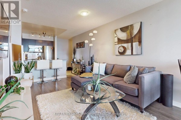 Living room with grey sectional sofa, glass coffee table, and open-concept kitchen area. - 1711 - 50 Lynn Williams Street, Toronto