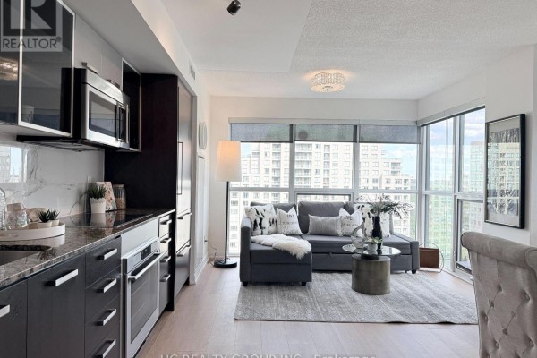 Kitchen with dark cabinets, stainless steel appliances, and open view to living room with gray sofa. - 1712 - 5168 Yonge Street, Toronto