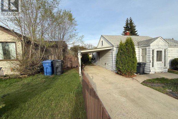 Exterior view of a white house with a carport, driveway, and surrounding green lawn. - 1712 103rd Street, North Battleford