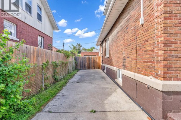 Concrete driveway runs alongside a brick house with a chain-link fence and wooden gate. - 172 Giles Boulevard, Windsor