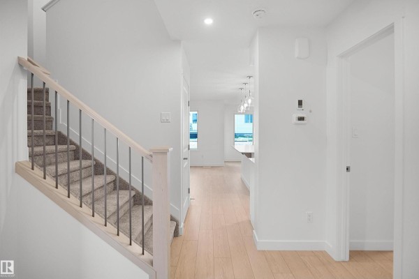 Interior hallway with light wood floors, white walls, carpeted stairs with metal railings, and a doorway to the kitchen. - 1721 8 St Nw, Edmonton