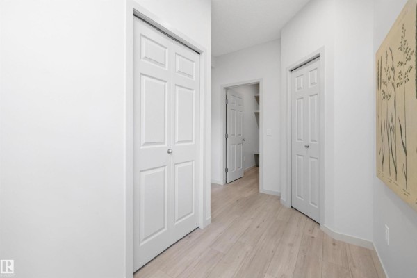 Interior hallway with light wood flooring, white walls, and multiple closet doors, showing an opening to another room. - 17431 7 St Ne, Edmonton