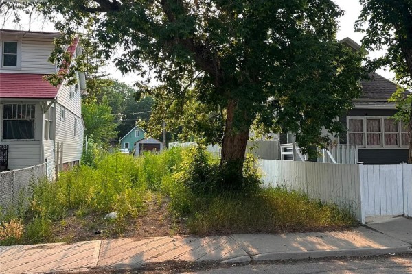View of a vacant lot with tall grass and a mature tree in the center, bordered by houses and fences. - 1760 Toronto Street, Regina