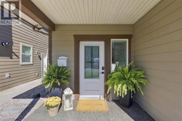 Entrance to the house with a white door, flanked by potted ferns and a welcome mat. - 1765 Foxtail Drive, Kamloops