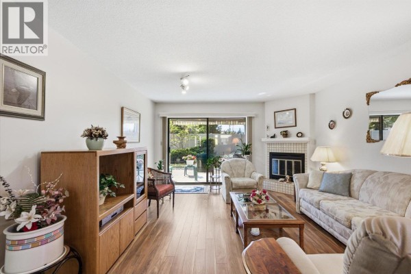 Living room with a fireplace, sofa, chairs, wooden entertainment unit, and a sliding glass door to the patio. - 1765 Leckie Road Unit# 201, Kelowna