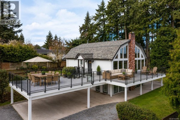 Rear view of the house with a spacious deck, garden beds, and trees. - 1766 Pritchard Rd, Cowichan Bay